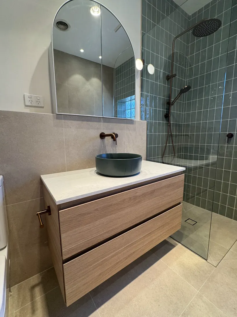 Sage green tiled bathroom with an arched mirror, oak vanity and a deep green basin